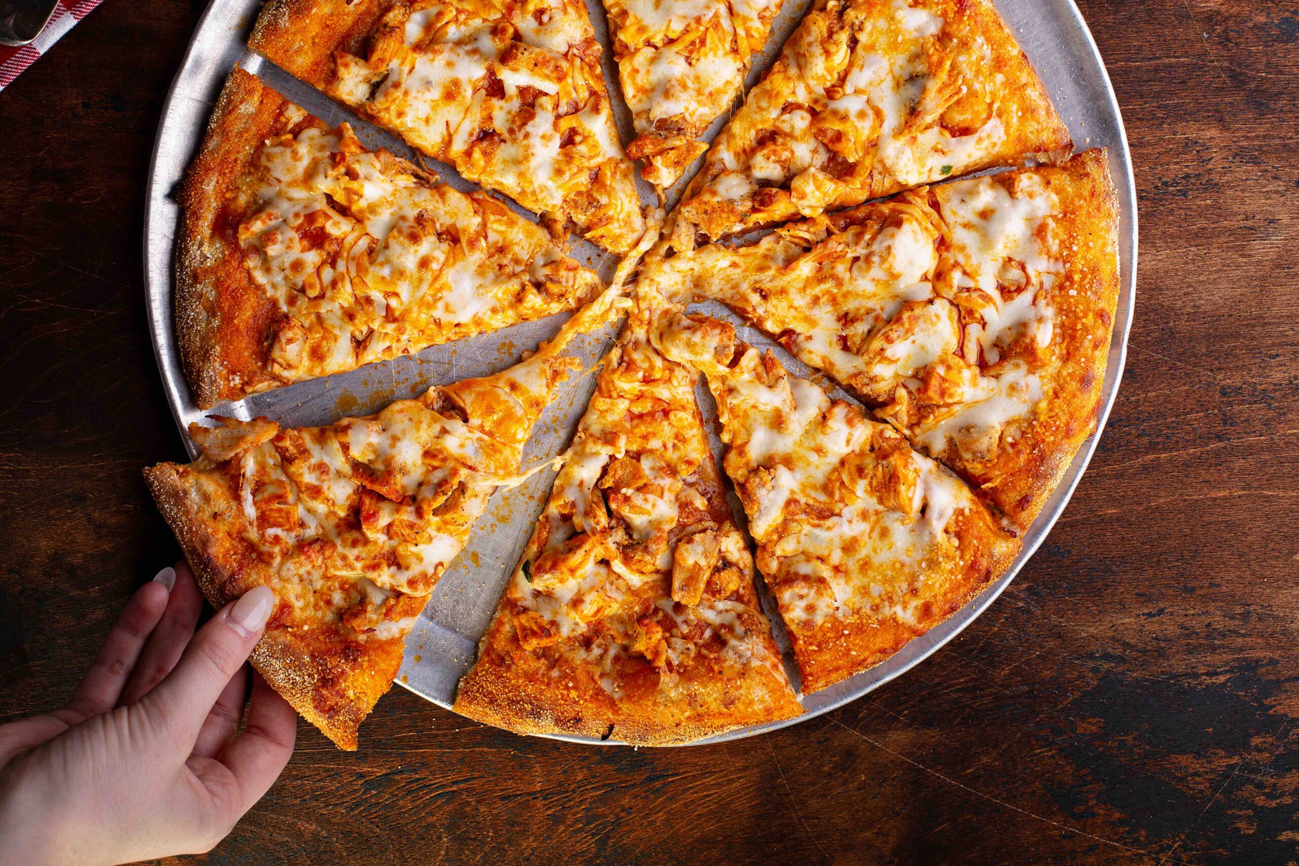 A hand takes a slice from a round pizza with melted cheese and toppings, served on a metal tray on a wooden table at Java House of Pizza.