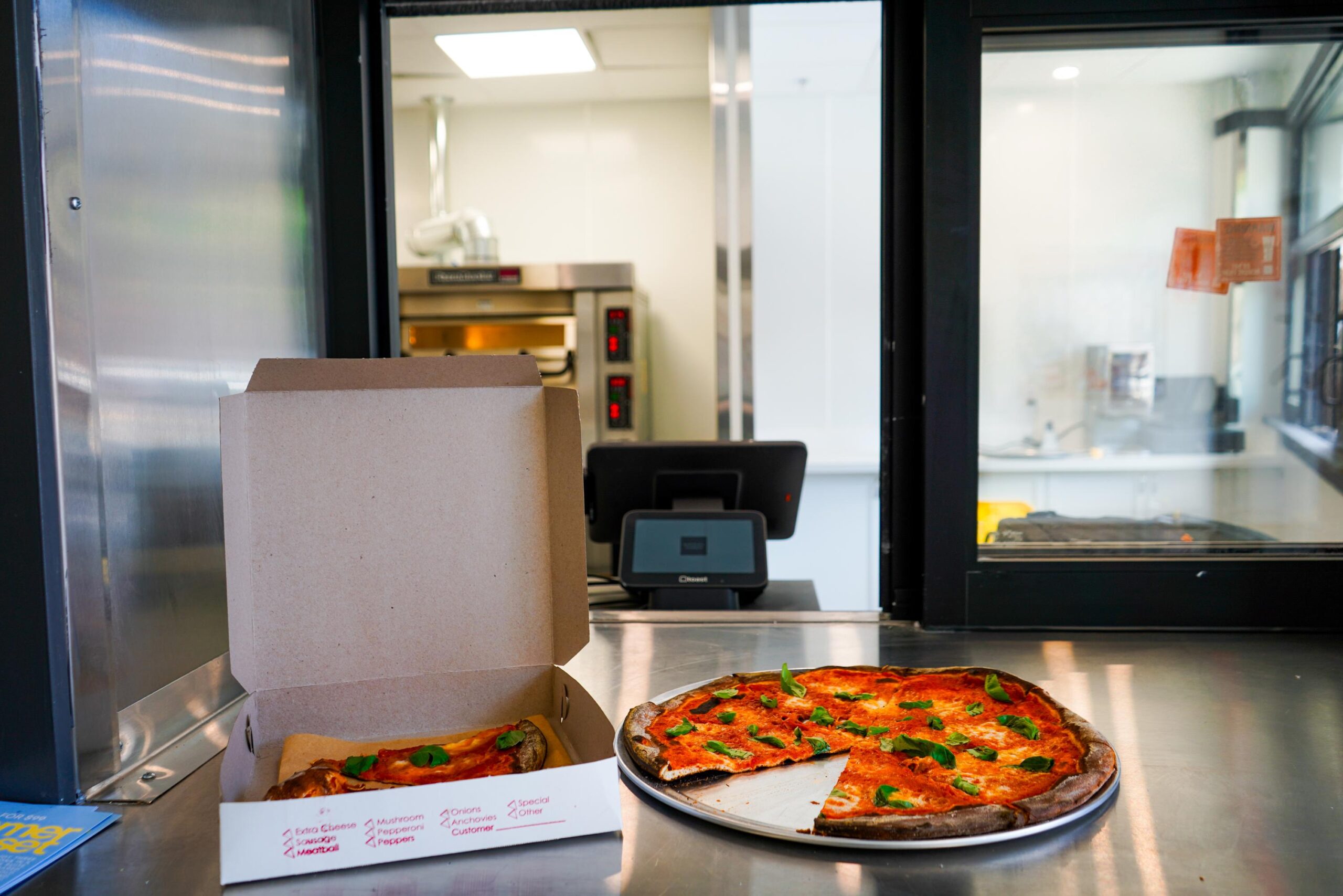 A pizza in a cardboard box sits next to a partially sliced pizza on a tray, both on a stainless steel counter in front of a kitchen window.