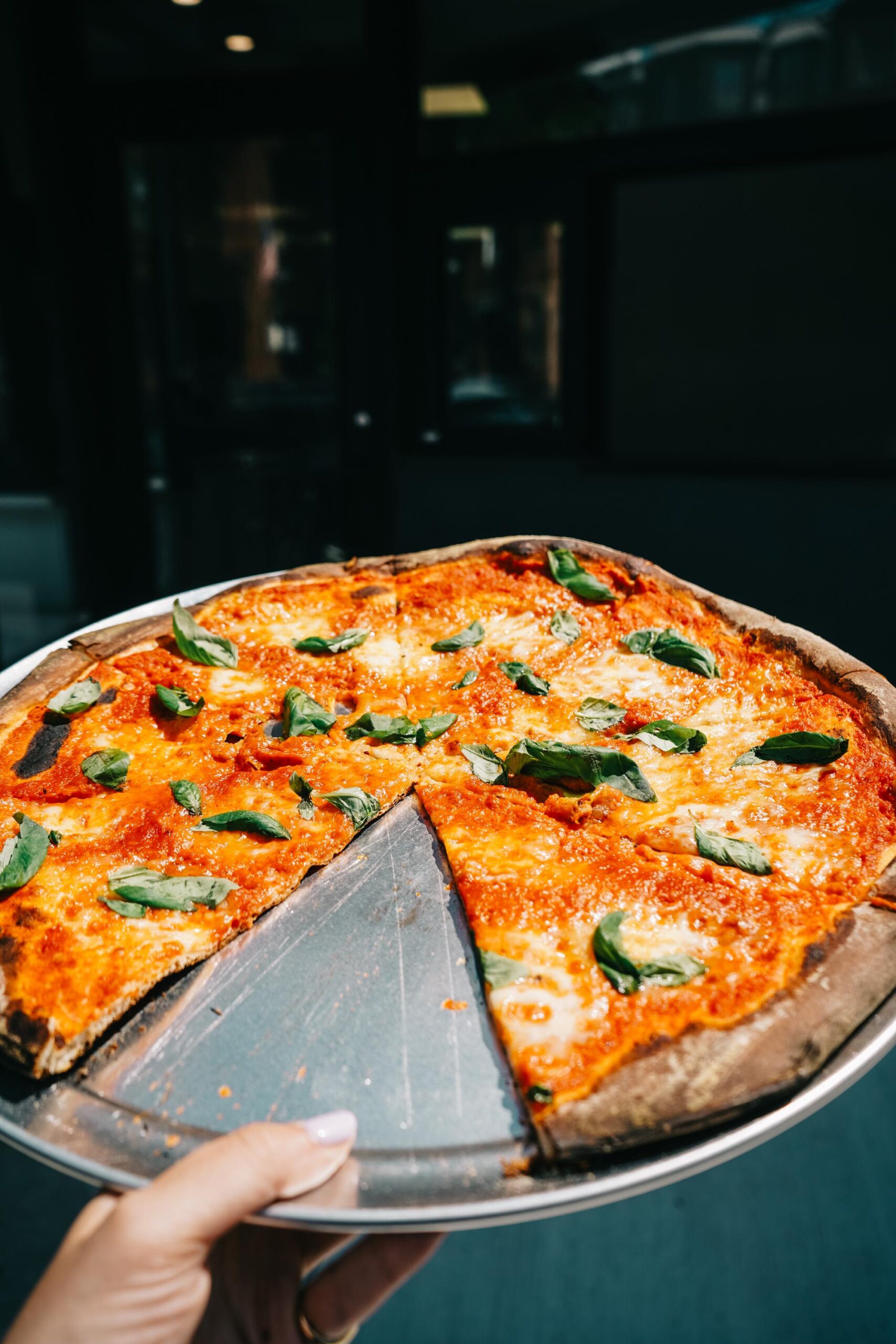 A hand holds a metal tray with a margherita pizza, missing one slice, topped with basil leaves, cheese, and tomato sauce.