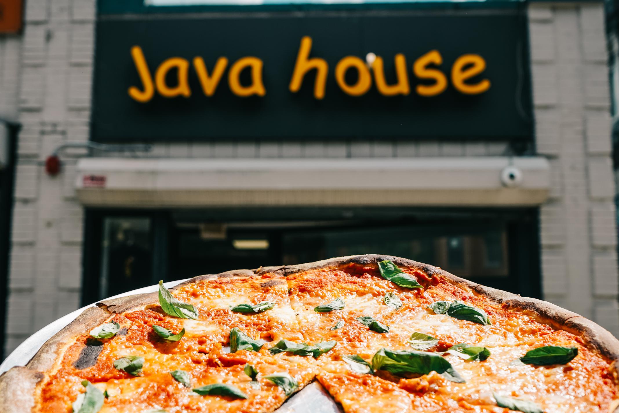 A margherita pizza with basil leaves in the foreground, with a sign reading 