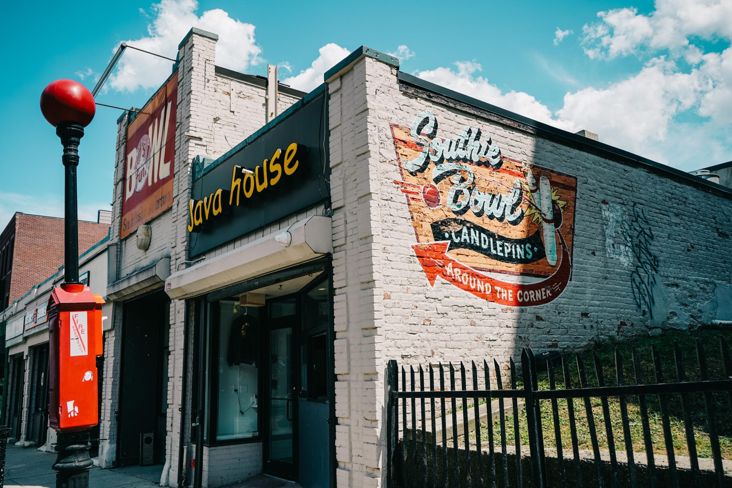 Street view of a beige brick building with signs reading “Java House” and “Southie Bowl Candlepins.” A red emergency call box and black metal fence are visible in the foreground.