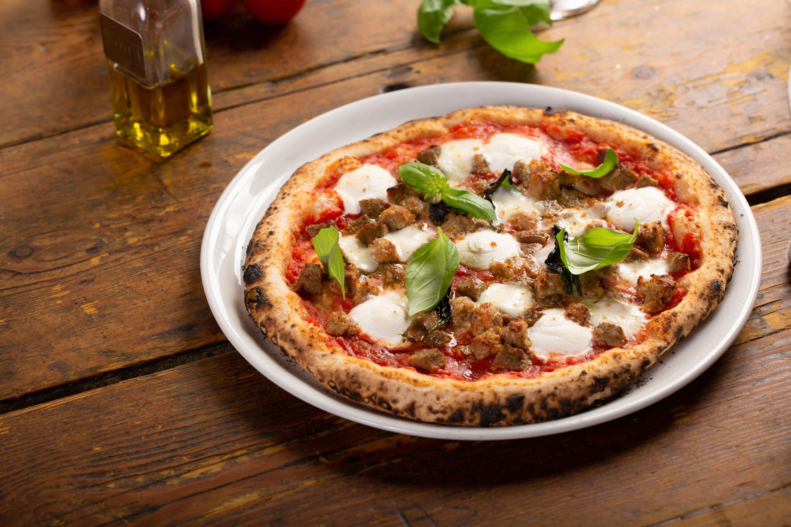 A Neapolitan-style pizza from Java House of Pizza sits on a white plate, topped with mozzarella cheese, sausage, tomato sauce, and fresh basil leaves, arranged beautifully on a wooden table.
